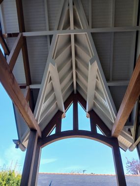 Vue en contre-plongée d'un pignon de porche décoratif en bois, avec des poutres en bois sombre et des panneaux de toit peints en blanc, sur fond de ciel bleu.