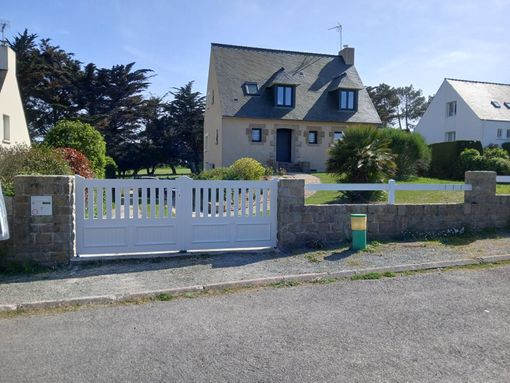 Une maison beige à deux étages, avec un toit foncé et des lucarnes, derrière une clôture et un portail blancs, par une journée ensoleillée.