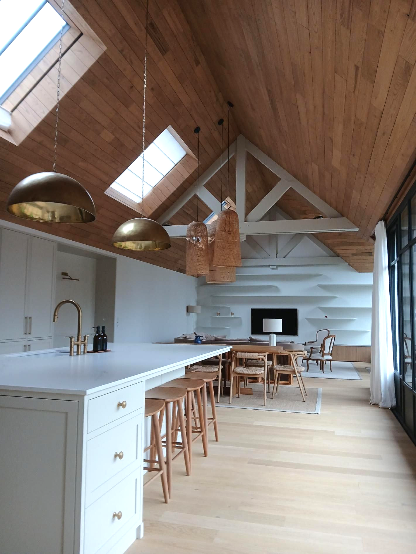 Une cuisine et un séjour modernes et ouverts, avec plafonds voûtés en bois, puits de lumière, luminaires en laiton et îlot central blanc.