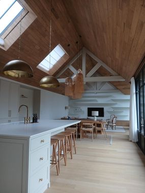 Une cuisine et un espace de vie lumineux et ouverts, avec de hauts plafonds lambrissés, des puits de lumière, des suspensions dorées et des armoires blanches.