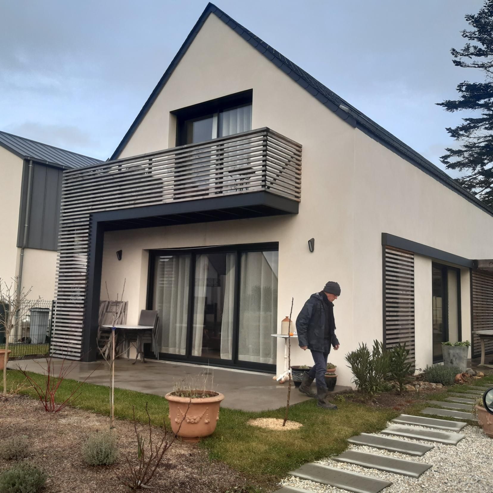 Une maison moderne aux tons clairs, avec des accents de lattes de bois, un balcon et des portes coulissantes, avec une personne marchant sur la pelouse.