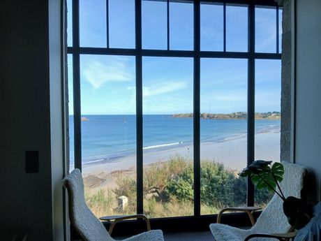 Deux fauteuils font face à une grande fenêtre donnant sur une plage et l'océan sous un ciel dégagé.