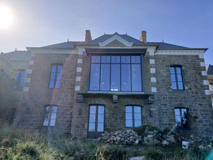 Une maison en pierre de deux étages avec une imposante extension centrale vitrée au deuxième étage, sous un ciel bleu.