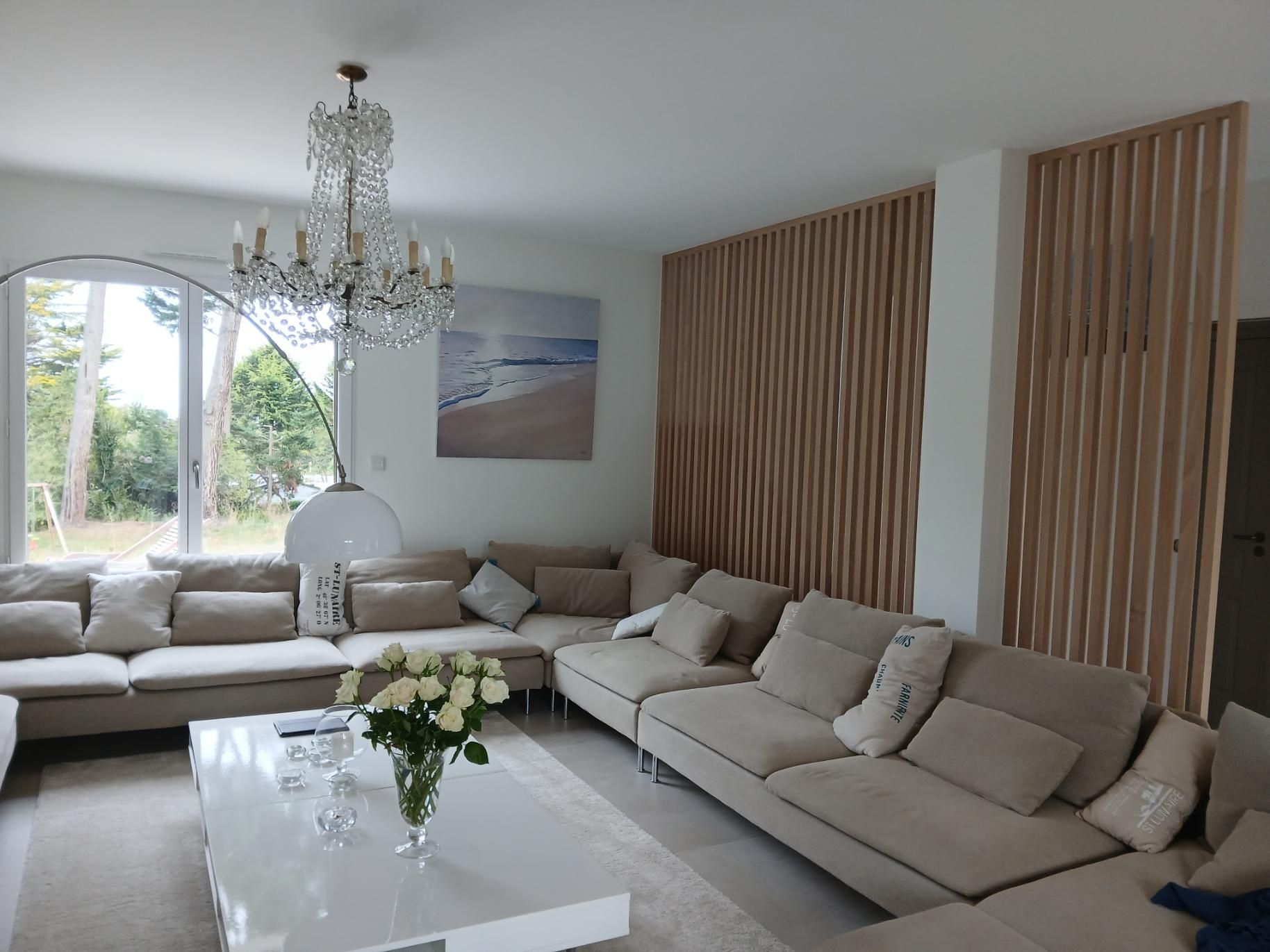Un salon confortable avec un grand canapé beige en forme de L, un lustre en cristal, une table basse blanche ornée de fleurs et des lattes de bois.