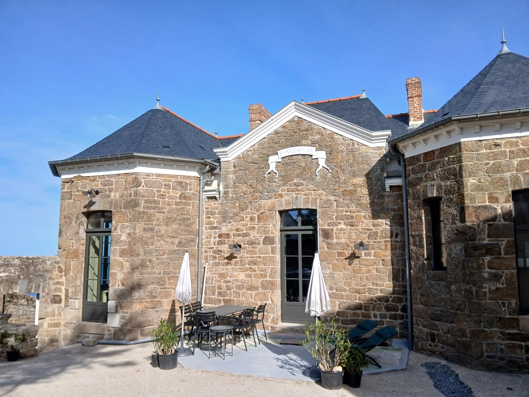 Une maison en pierre avec un toit en ardoise et un patio avec une table et des chaises sous deux parasols par une journée ensoleillée.