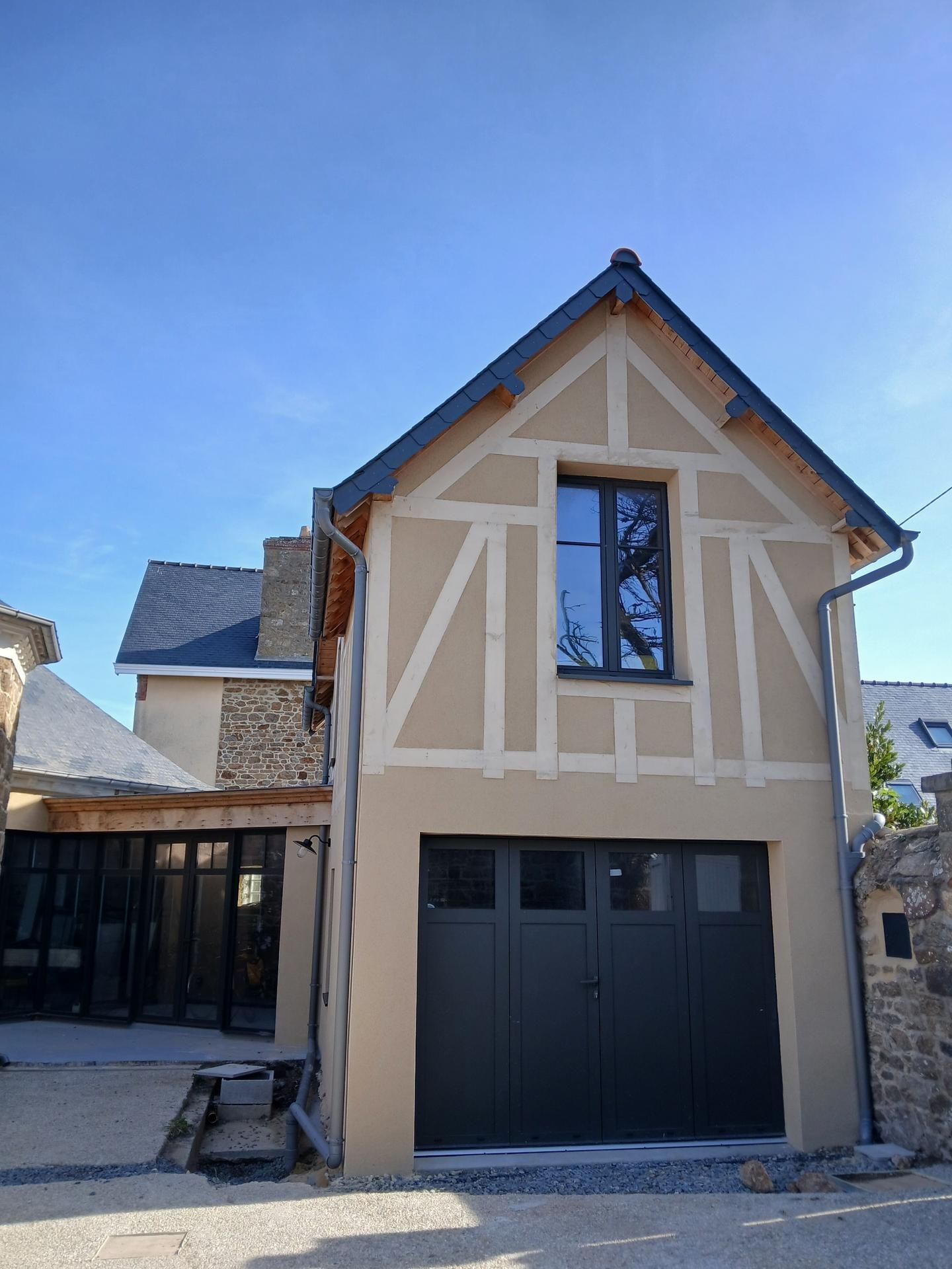 Un bâtiment à colombages de deux étages, aux murs beiges et aux boiseries gris foncé, comportant une porte de garage et une fenêtre à l'étage.