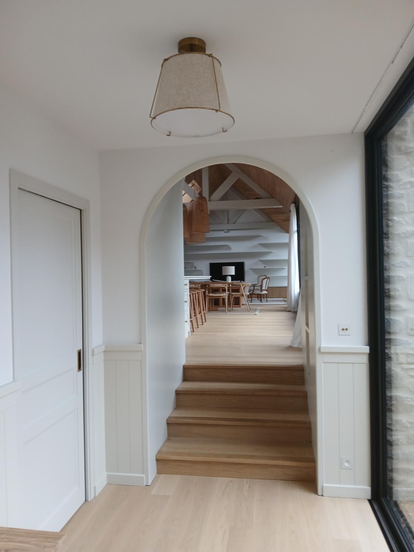 Un couloir clair avec un escalier en bois menant, par une porte cintrée, à une pièce rustique aux poutres apparentes.