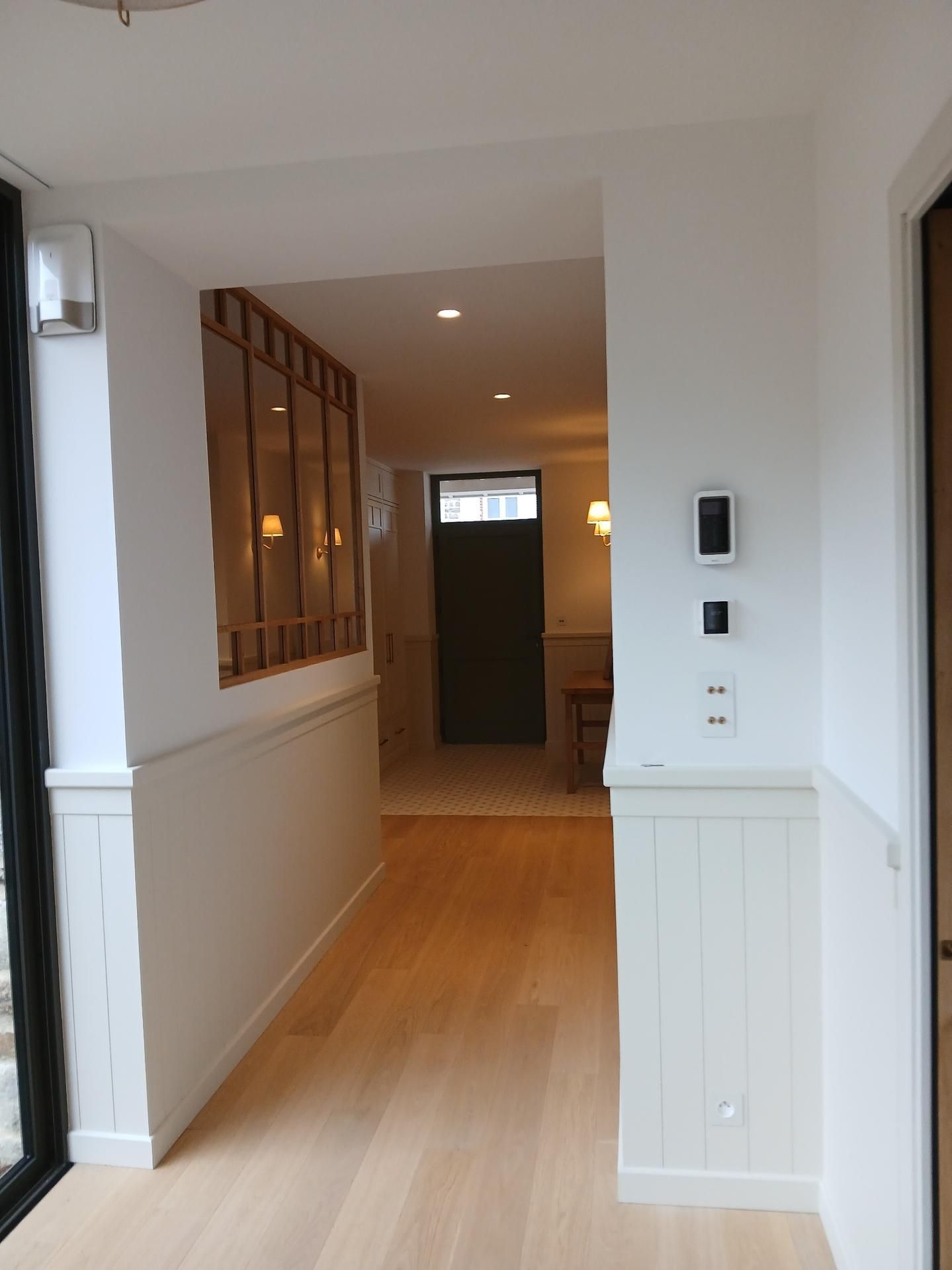 Un couloir avec un parquet clair et des lambris blancs menant à une porte, avec une fenêtre intérieure sur la gauche.