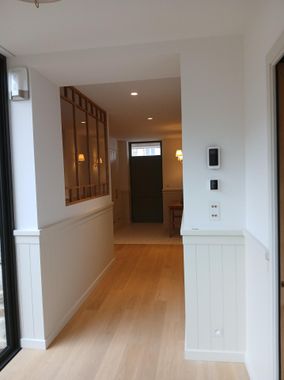Un couloir avec un parquet clair et des lambris blancs menant à une porte, avec une fenêtre intérieure sur la gauche.