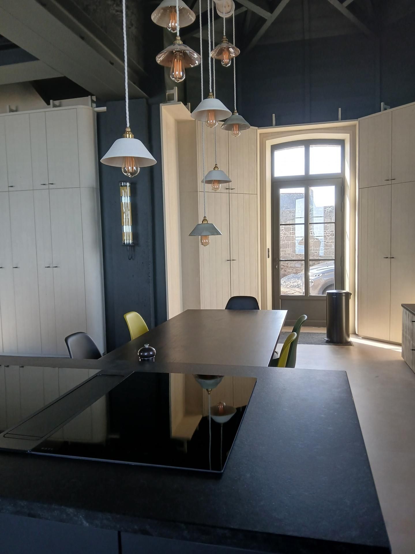 Un îlot de cuisine sombre, équipé d'une plaque de cuisson, met en valeur une table à manger éclairée par un ensemble de suspensions lumineuses de style industriel.