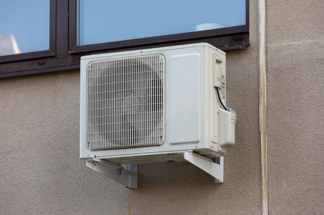 Unidad de aire acondicionado blanca instalada en la pared, debajo de una ventana en un muro exterior beige