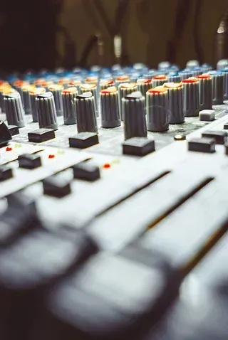 Consola de mezcla de sonido con perillas y deslizadores, enfocada en los controles, poco iluminada.