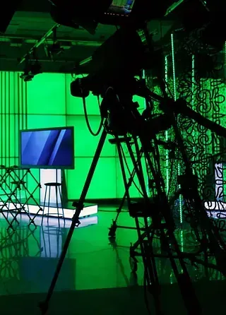 Silueta de una cámara de televisión sobre un trípode en un estudio de pantalla verde. Televisión y taburete al fondo.