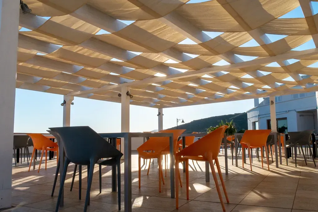 Comedor al aire libre con sillas naranjas y grises alrededor de mesas, bajo un toldo de tela beige. La luz del sol brilla a través de él.