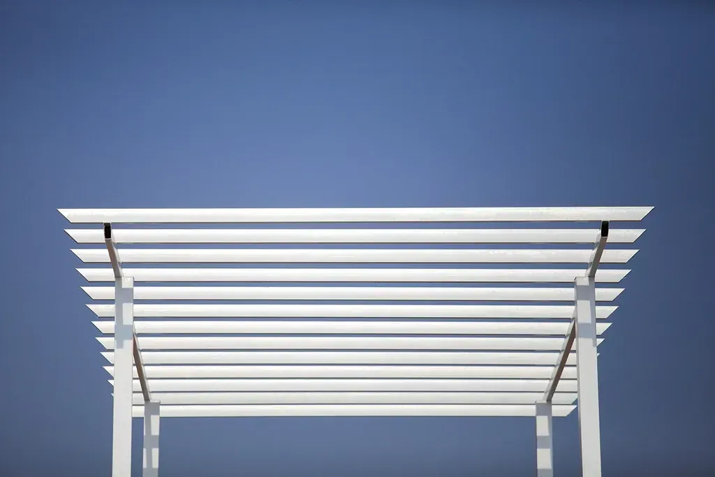 Pérgola blanca contra un cielo azul claro. Lamas horizontales que proyectan sombras.