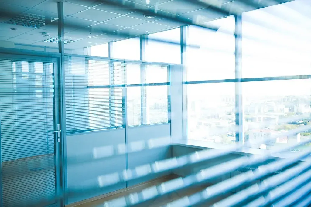 Vista interior de una oficina moderna con grandes ventanas y persianas que proyectan una suave luz azul.