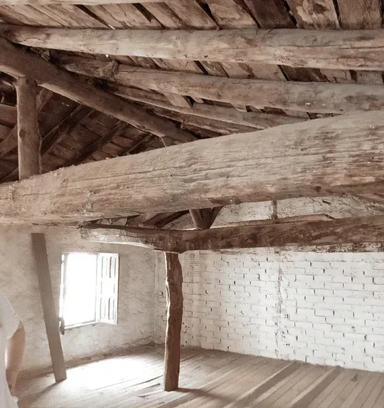 Vigas de madera en el techo y postes de soporte en una habitación rústica. Una ventana con contraventanas deja entrar la luz, realzando las paredes de ladrillo pintadas de blanco y el suelo de madera.