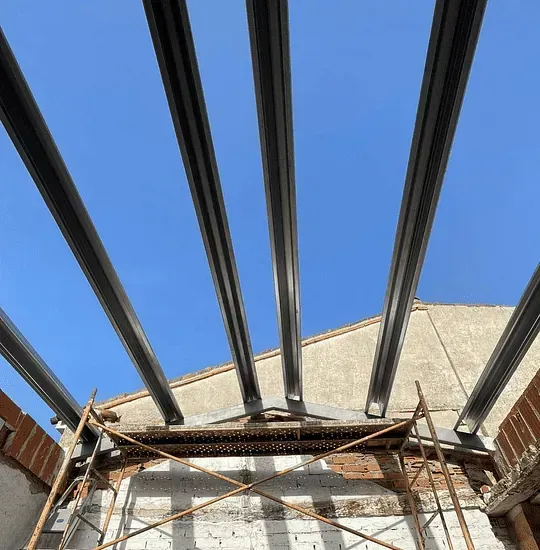 Vigas de acero que se extienden a través de un cielo azul sobre un edificio en construcción, sostenidas por andamios y ladrillos.