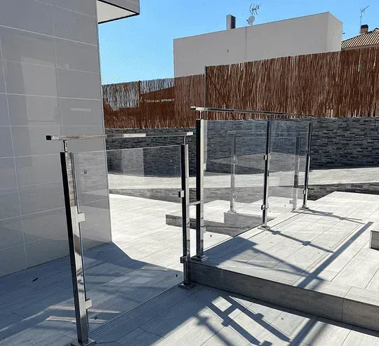 Un moderno espacio exterior con barandilla de vidrio y acero. Cuenta con terraza, baldosas y una cerca de bambú bajo un cielo azul.