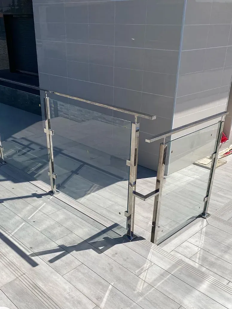 Una vista luminosa y soleada de la barandilla de vidrio y metal de un edificio moderno sobre una superficie pavimentada. El edificio es blanco y la barandilla proyecta largas sombras.