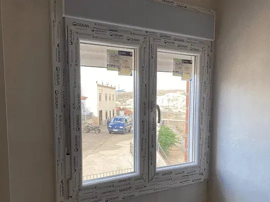 Ventana de doble acristalamiento con marcos blancos. A través del cristal se ve una calle con edificios y un coche azul.
