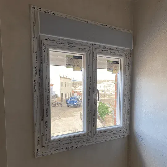 Ventana con marco blanco y persiana enrollable, que muestra una vista de una calle con edificios y un automóvil azul.