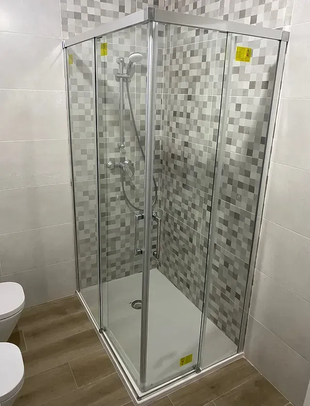 Mampara de ducha esquinera con puerta de cristal, marco plateado y base blanca. La ducha tiene un rociador de pared y está revestida con un diseño de mosaico.