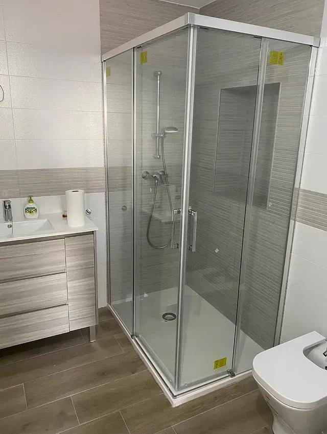 Baño moderno con mampara de ducha de cristal, lavabo, inodoro y suelo imitación madera clara. Las paredes tienen azulejos texturizados de color claro.