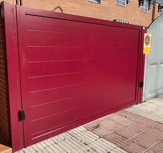 Puerta metálica roja con líneas horizontales, situada en una acera de ladrillo frente a un edificio. Se ven un letrero amarillo y una puerta de color más claro.