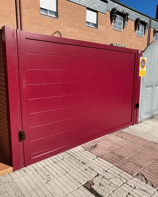 Puerta metálica roja y horizontal con líneas horizontales sobre una acera de ladrillo. Detrás hay un edificio con puerta y ventanas grises.