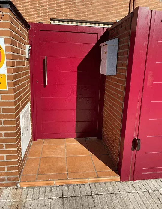 Puerta metálica roja y buzón en una pared de ladrillo. La entrada tiene azulejos marrones.