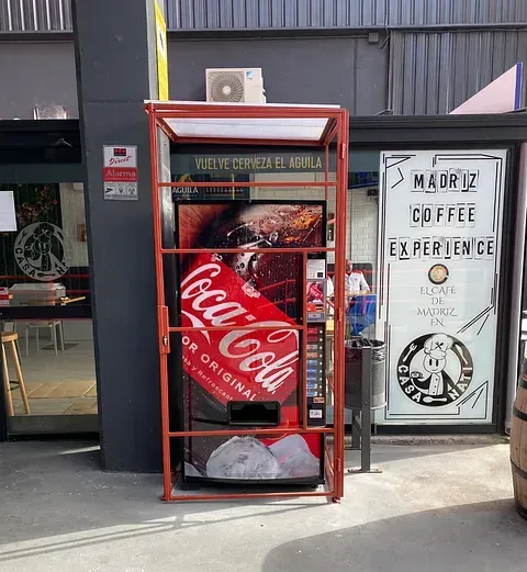 Máquina expendedora roja de Coca-Cola con una jaula protectora, frente a un negocio. Un cartel junto a ella anuncia una