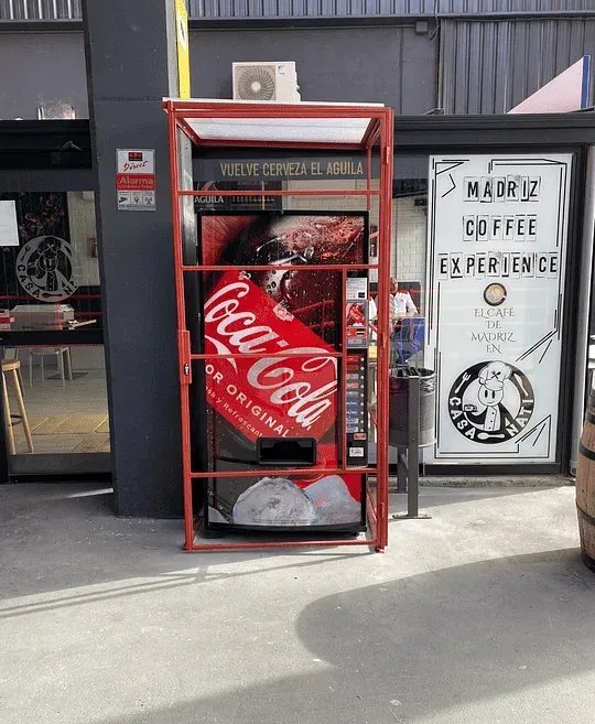 Afuera, hay una máquina expendedora de Coca-Cola con una estructura metálica roja. Junto a ella, un cartel anuncia «Madriz Coffee Experience».