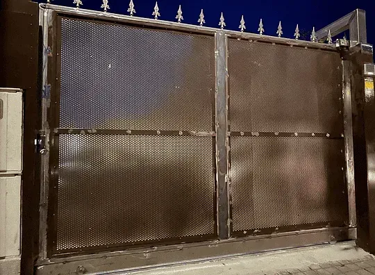Puerta metálica oscura y cerrada con malla romboidal. Tiene una parte superior con púas y está ambientada en un ambiente nocturno.