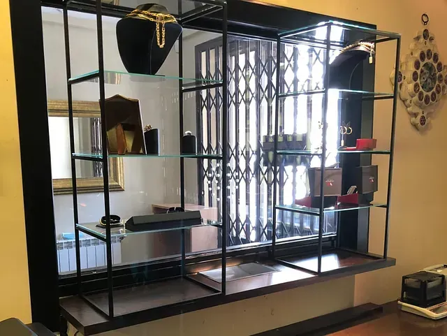 Vitrina con joyas sobre estantes de cristal, frente a una ventana con reja de seguridad. Los estantes y el marco son negros, y las paredes amarillas.