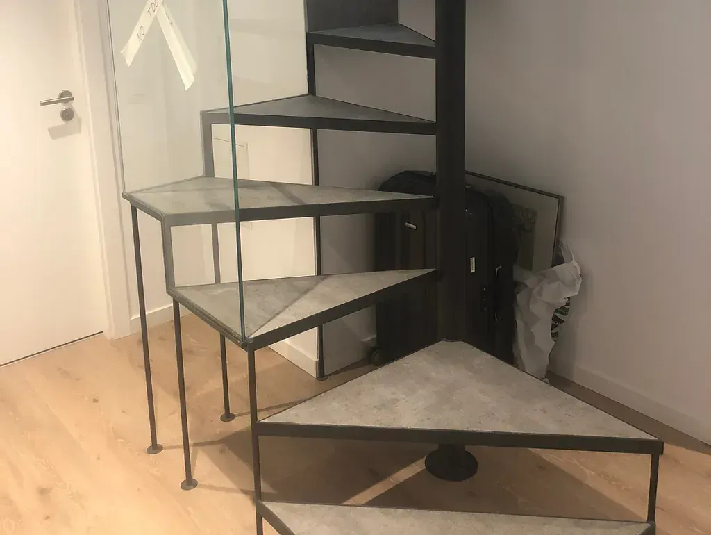Escalera de caracol triangular de metal contra una pared blanca. Al fondo, se aprecian suelos de madera clara y una puerta parcialmente visible.