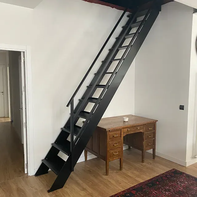 Escalera de metal negro que sube a un altillo; junto a un escritorio de madera. Paredes blancas, suelo de madera.