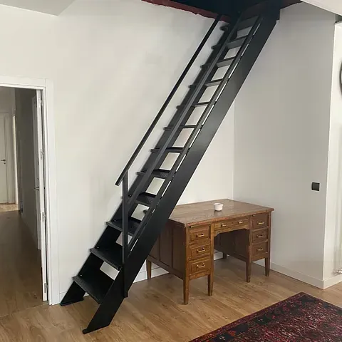 Escalera de metal negro que conduce a un loft en una habitación con un escritorio de madera y paredes blancas.