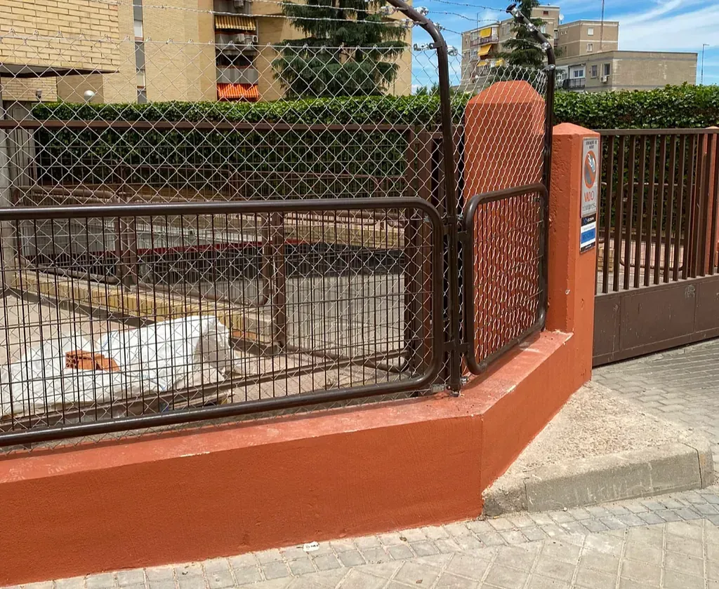 Una valla de ladrillo y metal marrón rodea una obra en construcción; una base de pared de color naranja y una acera en primer plano.