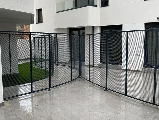 Una valla metálica negra rodea un patio y encierra parcialmente un pequeño jardín. Edificio blanco al fondo.