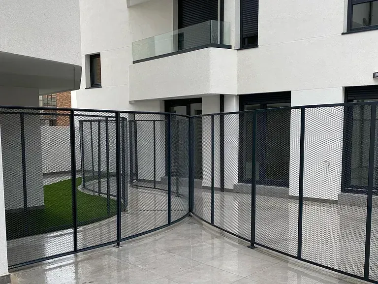 Valla metálica negra que delimita una zona exterior con césped artificial. La valla delimita un edificio con balcón y ventanas oscuras.