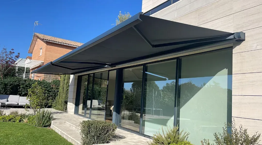 Un toldo retráctil gris oscuro que da sombra a un edificio con paredes de vidrio en un día soleado.