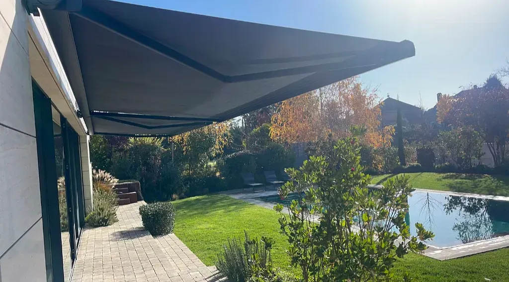 Un toldo gris que se extiende desde un edificio con un patio de ladrillo con vista a un patio trasero con piscina y árboles.