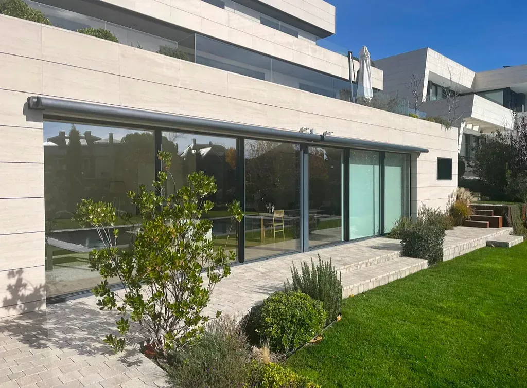Vista exterior de un edificio moderno con grandes ventanales, un patio de piedra y césped verde bajo un cielo azul.