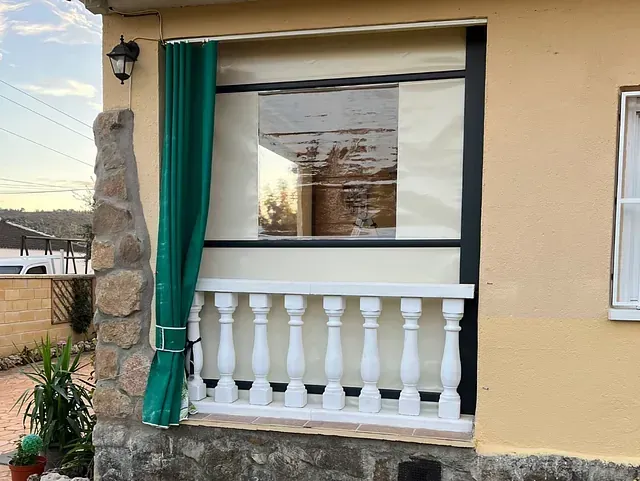 Vista exterior de una casa amarilla con ventana, cortina verde y balaustrada blanca. La ventana refleja una escena al aire libre.