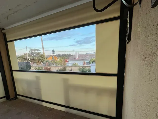 Una ventana parcialmente cubierta, con una persiana marrón bajada, con vistas a un barrio. El cielo es azul con algunas nubes.