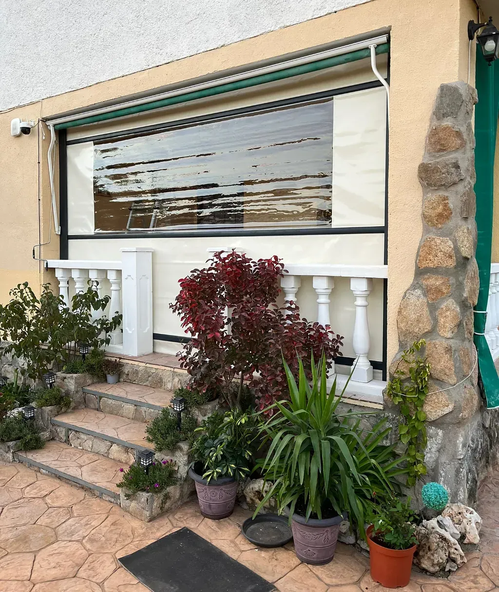 Entrada de una casa con escalones, plantas, un toldo retráctil y una columna de piedra. El toldo está desplegado, ocultando parcialmente la ventana.