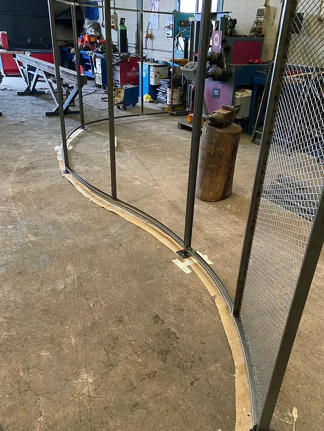 Una estructura metálica curvada con paneles de malla en un taller. La estructura está anclada al suelo de hormigón.