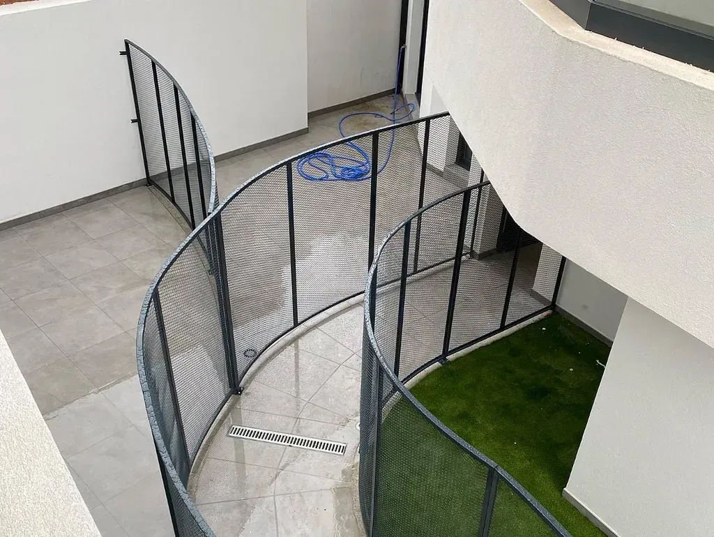 Una valla metálica negra curva encierra un sendero con una pequeña zona de césped artificial. El entorno es un edificio moderno de varias plantas.