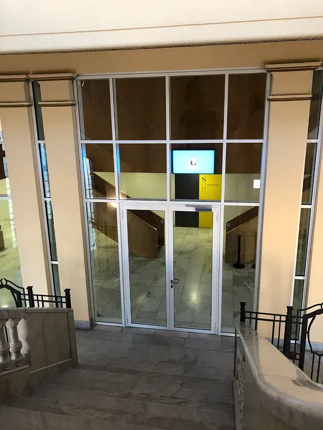 Puertas de cristal que dan a un vestíbulo con pantalla y escaleras. El ambiente es la entrada a un edificio de colores crema y tostado.